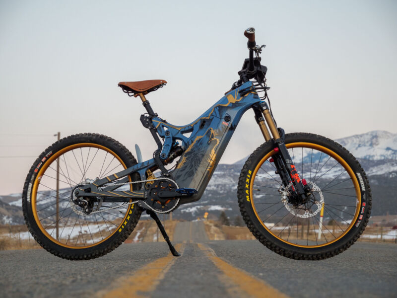 A wide view showing an Optibike E-Bike with snow covered mountains in the background, showing the hand painted frame featuring a wolf howling at the sky, painted in vibrant gold color.